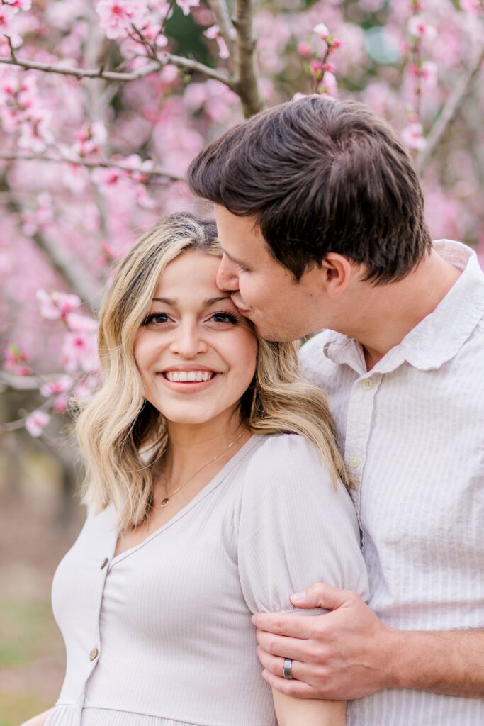 Soon to be mom smiles at the camera while soon to be dad kisses her forehead in a peach blossom orchard in Charlotte NC