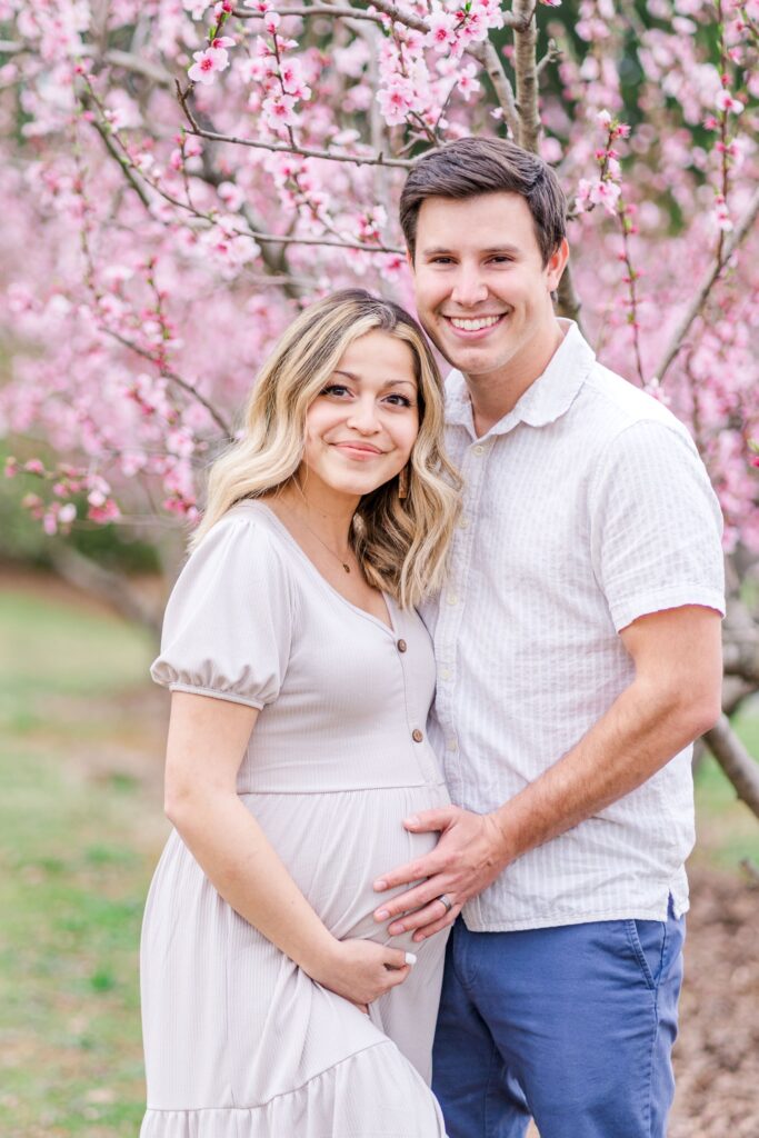 Soon to be parents smiling and posing in a peach blossom orchard in Charlotte NC