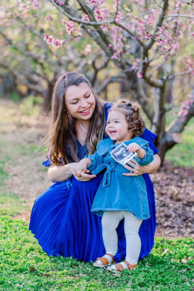 Expecting mom smiles at young daughter who is holding the sonogram of her brother. Both are smiling in a peach blossom orchard in Charlotte NC