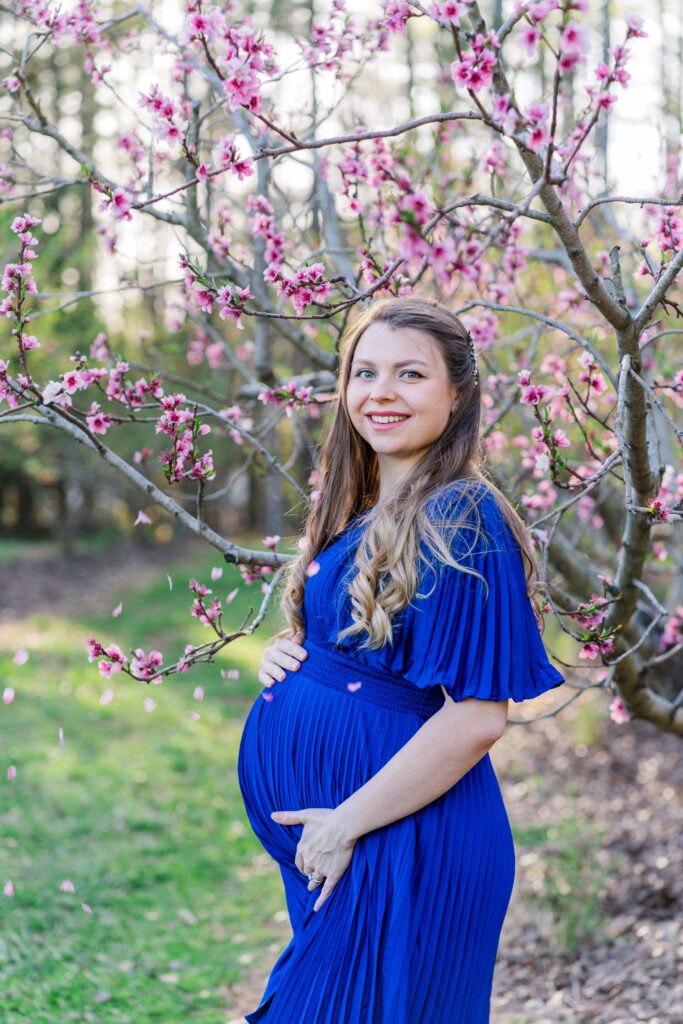 Expecting mother smiles in a peach orchard in Charlotte NC. Petals cascade down her blue maternity dress
