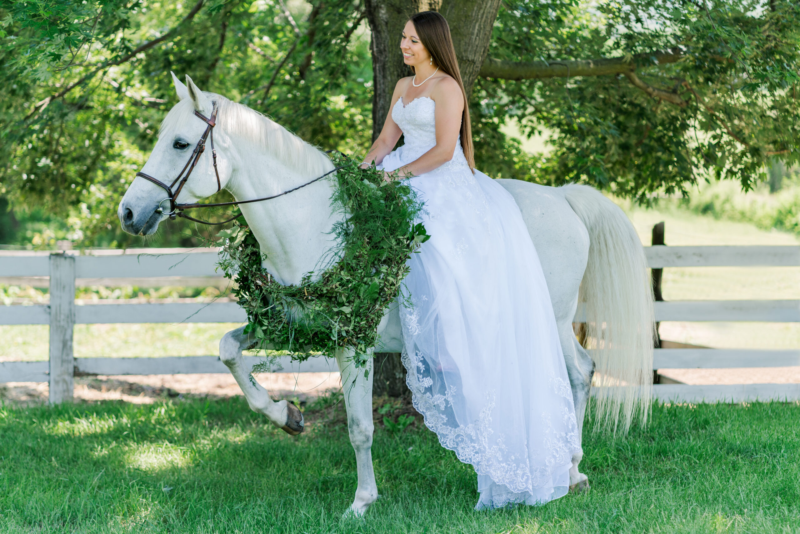 Kelli wears her wedding dress and sits on her white mare Lily in the green grass next to a white fence line