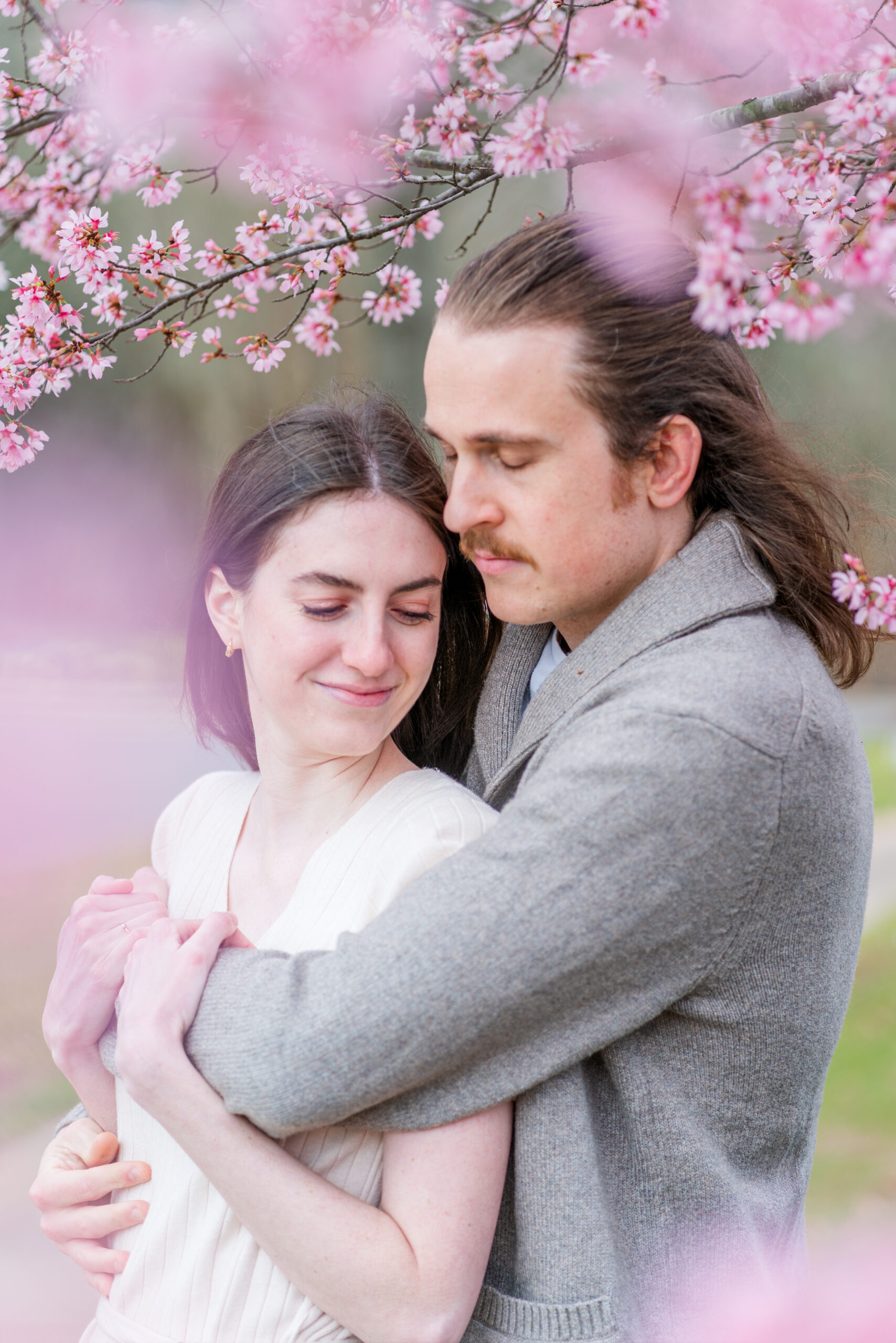 Couple Noelle and Hayden Hug each other amongst the pale pink blossoms of the cherry trees.