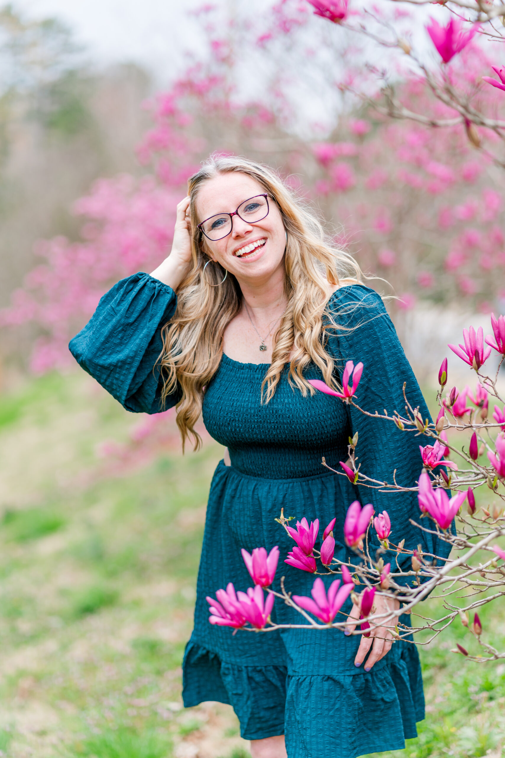 Stephanie smiles in a teal dress while standing in a "field" of bright pink tulip magnolias.