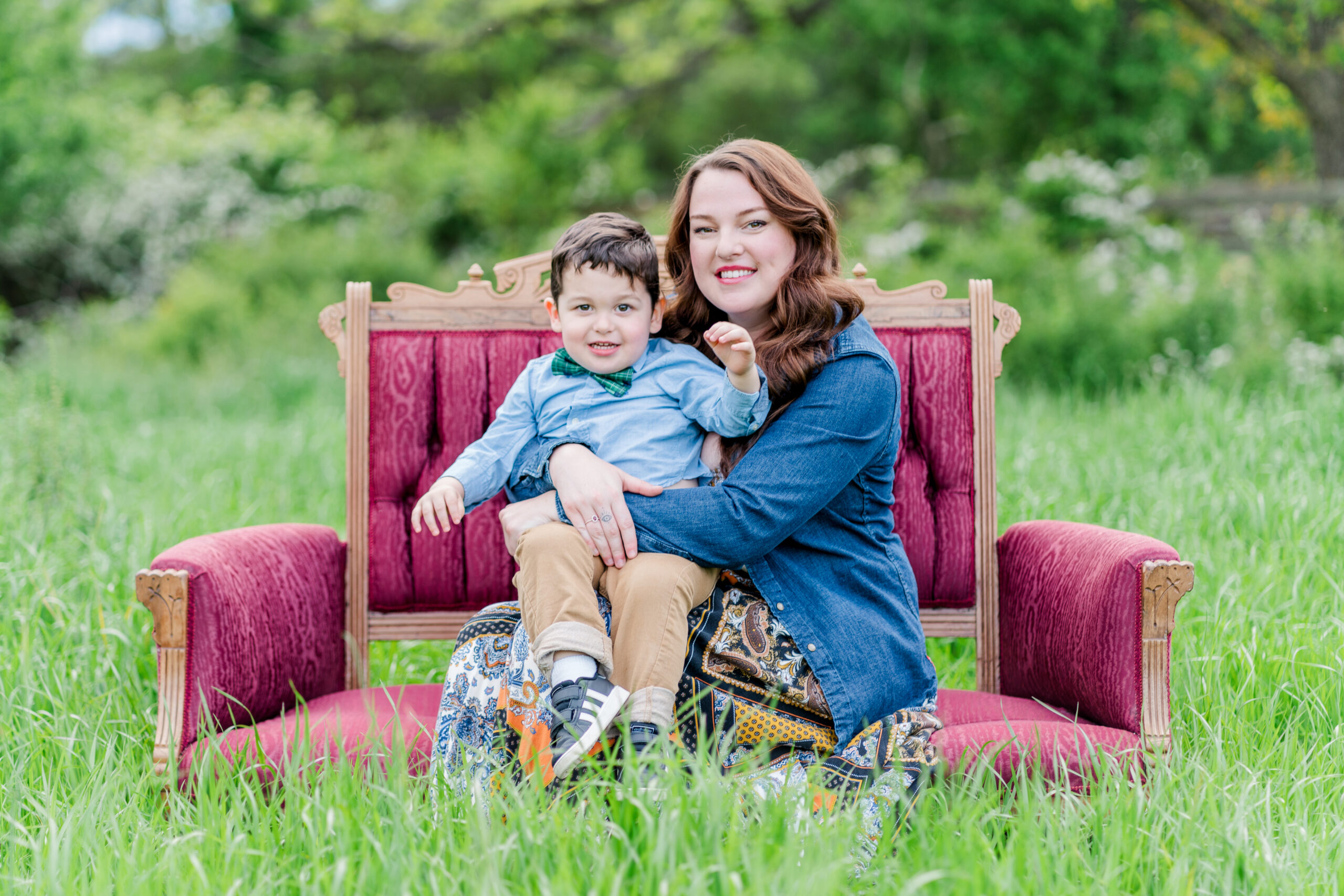 Mom Katy sits on a red vintage loveseat in a green field filled with tall grass. Her son Davis sits in her lap