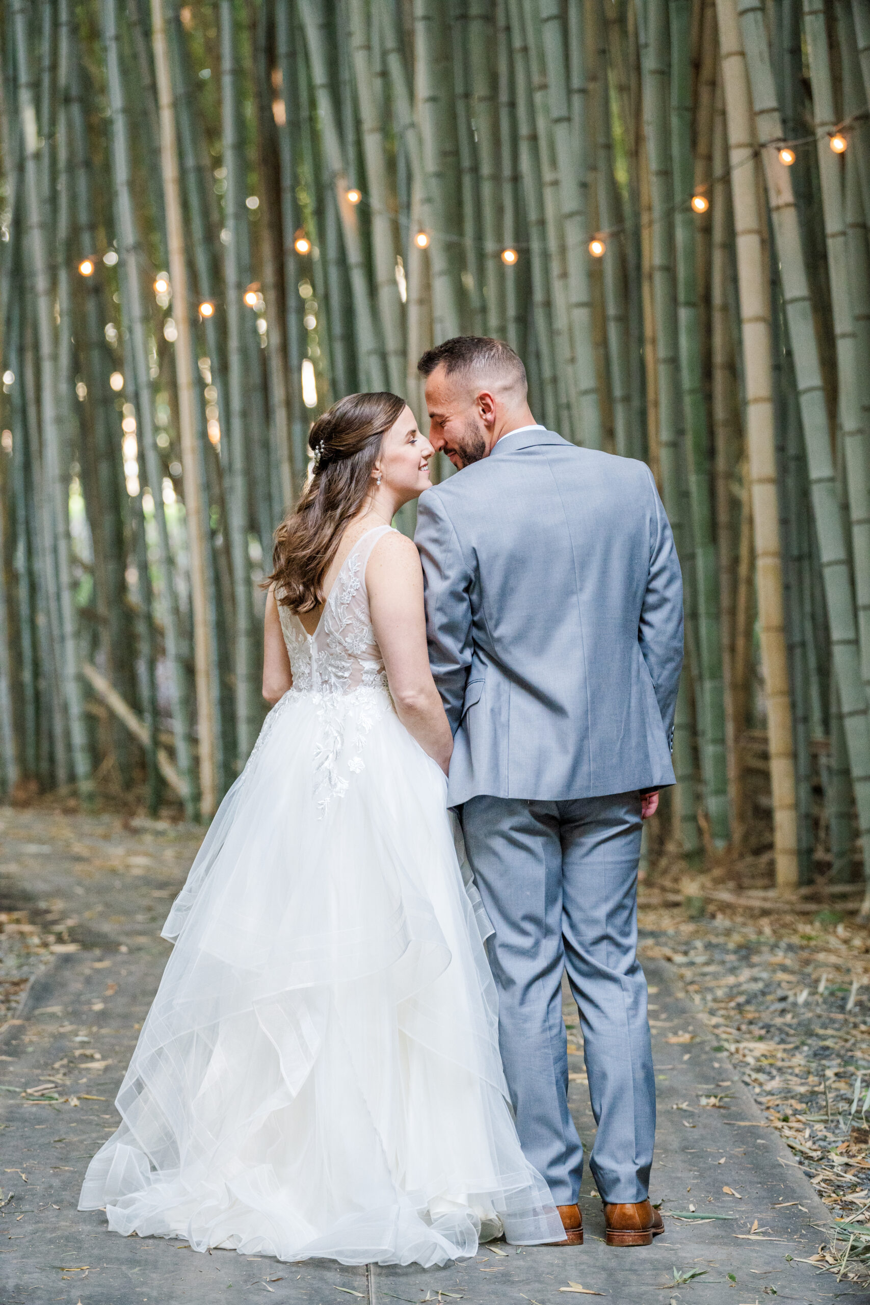 The bride and groom have their backs to the camera. Both their heads are turned in towards each other. Their eyes are closed as they smile and nuzzle in close. Around them is a towering forest of bamboo littered with dancing fairy lights.