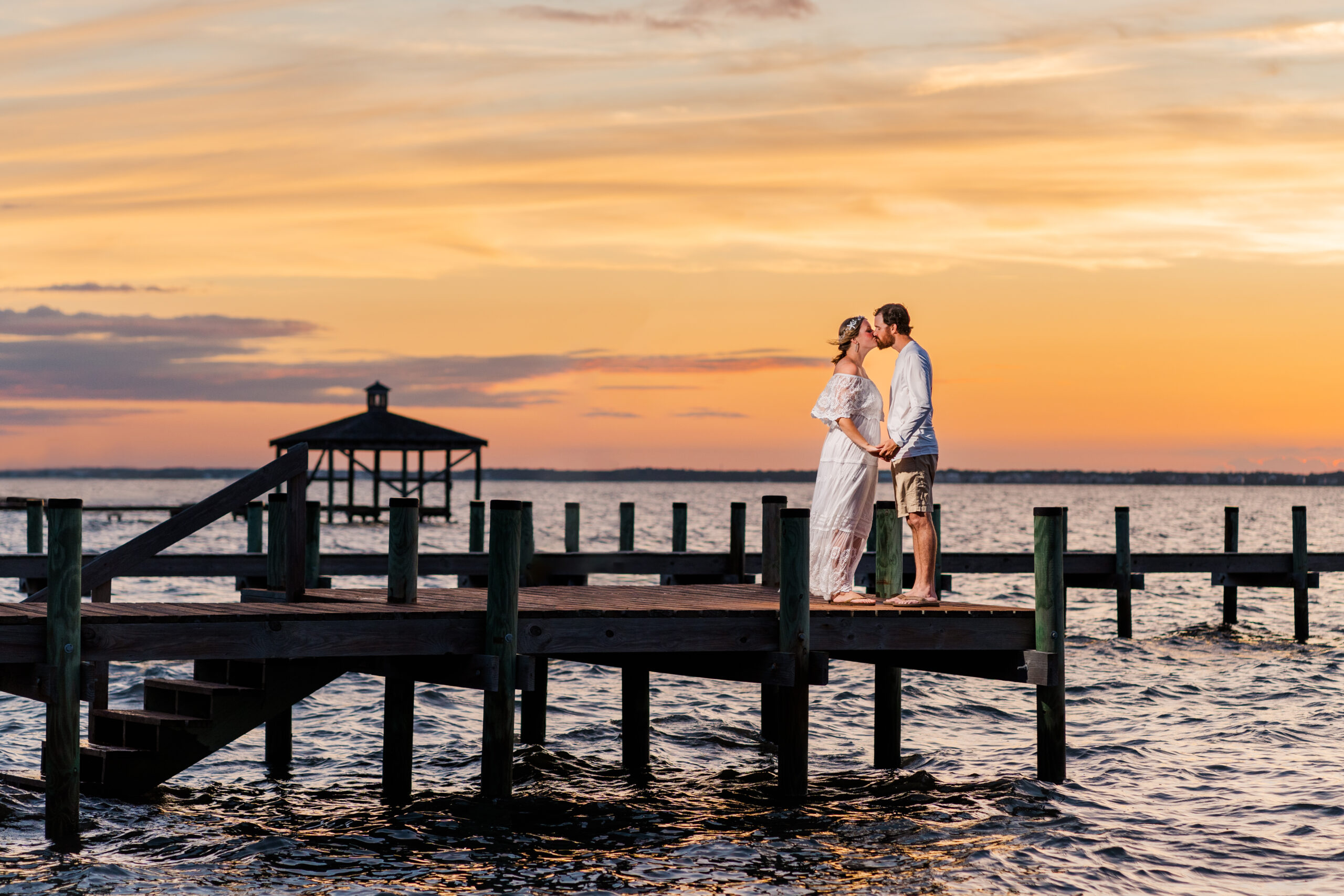 Katie and Jeffrey hold hands and kiss on a pier above the sound. The sunset blazes red, orange, and yellow behind them in the way that only beach sunsets can.