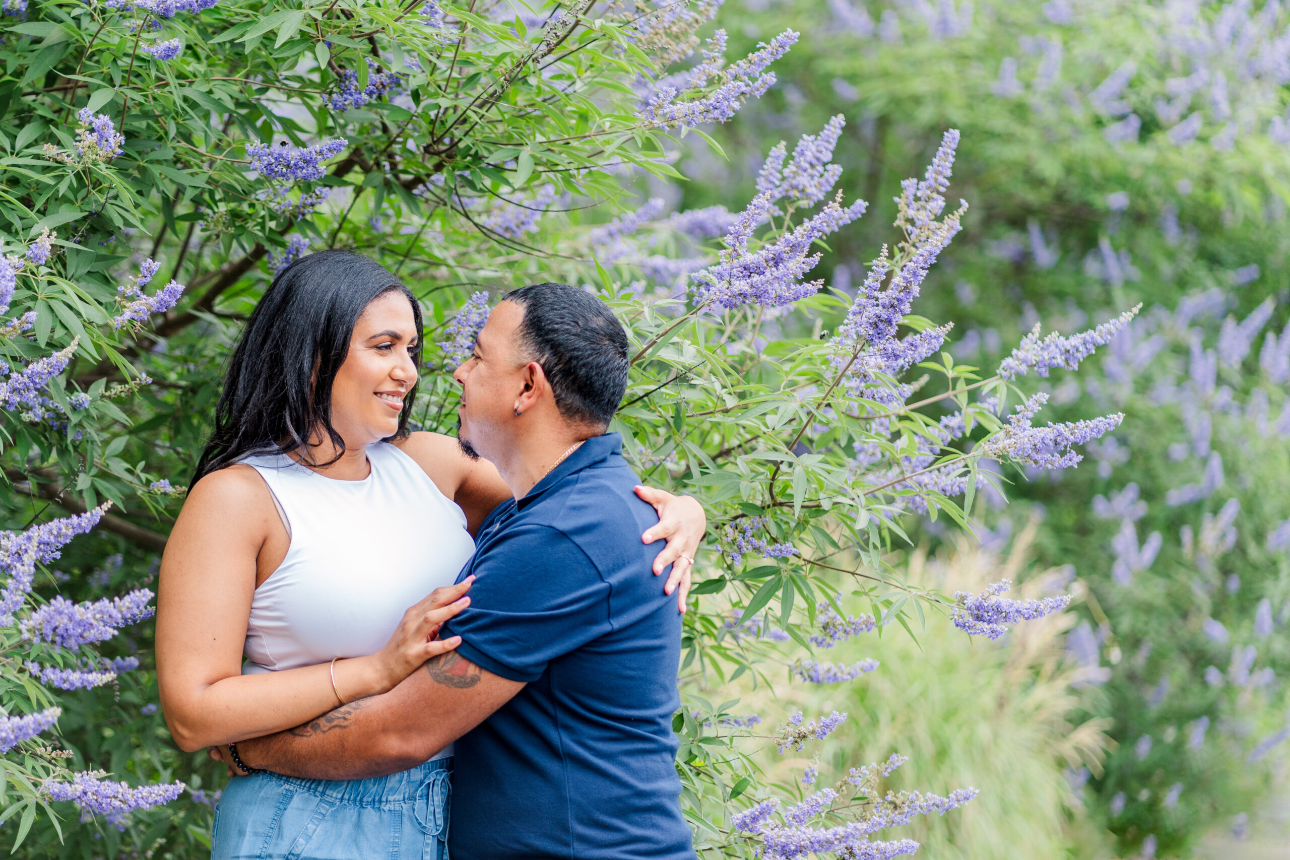 Zaida and Julio snuggle in close and look into each other's eyes. Both are nestled in a bush that contains a sea of purple flowers.