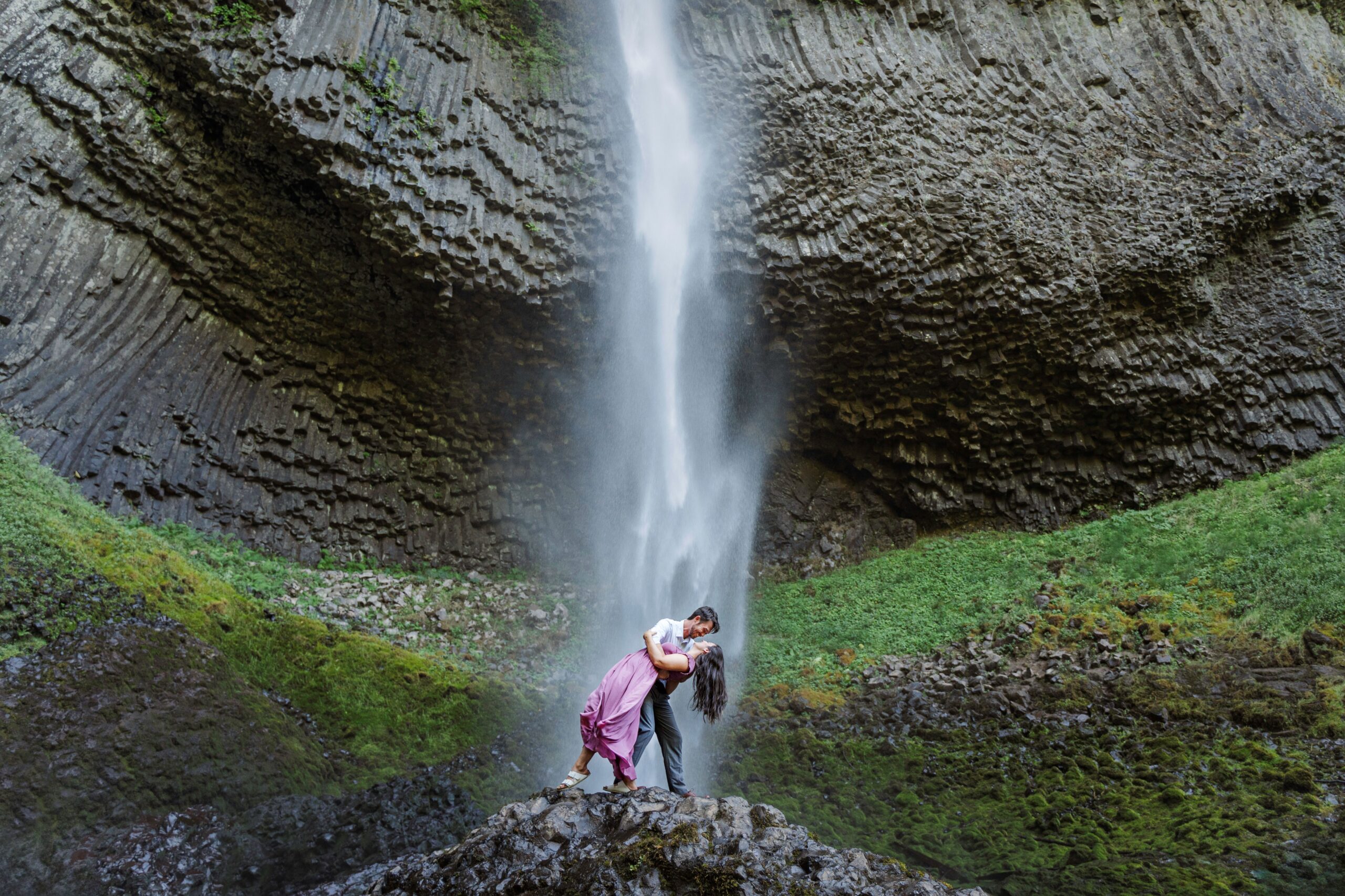 Aaron and Delanie stand on rock beneath the waterfall. Latourell Falls, a long waterfall surrounded by moss and dark basalt columns, rages behind them as Aaron dips Delanie down for a kiss.