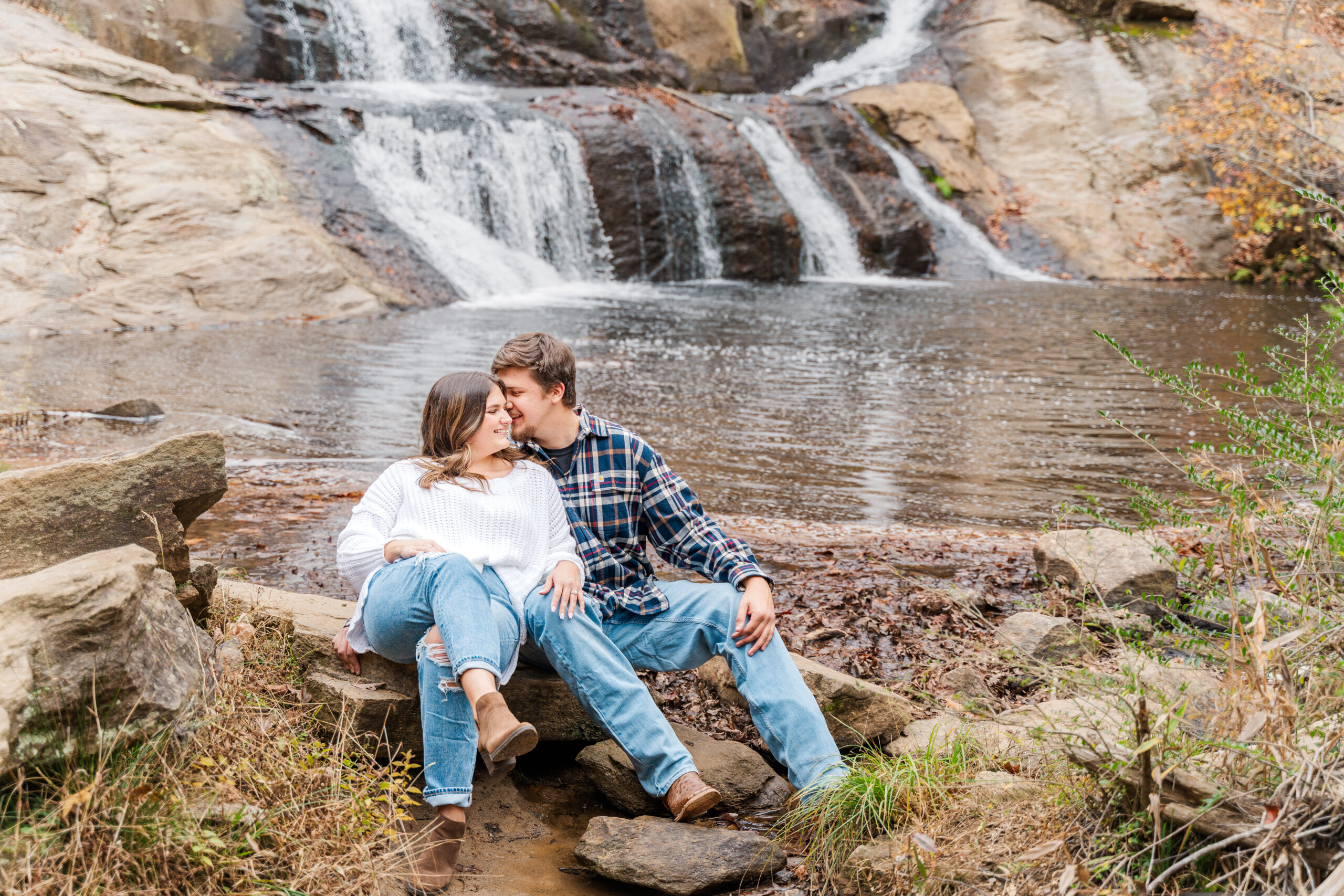 Newly engaged couple Cheyenne and Ben snuggle up close together on a rock on the bank of small pool. A beautiful two-tiered waterfall flows in the background behind them.