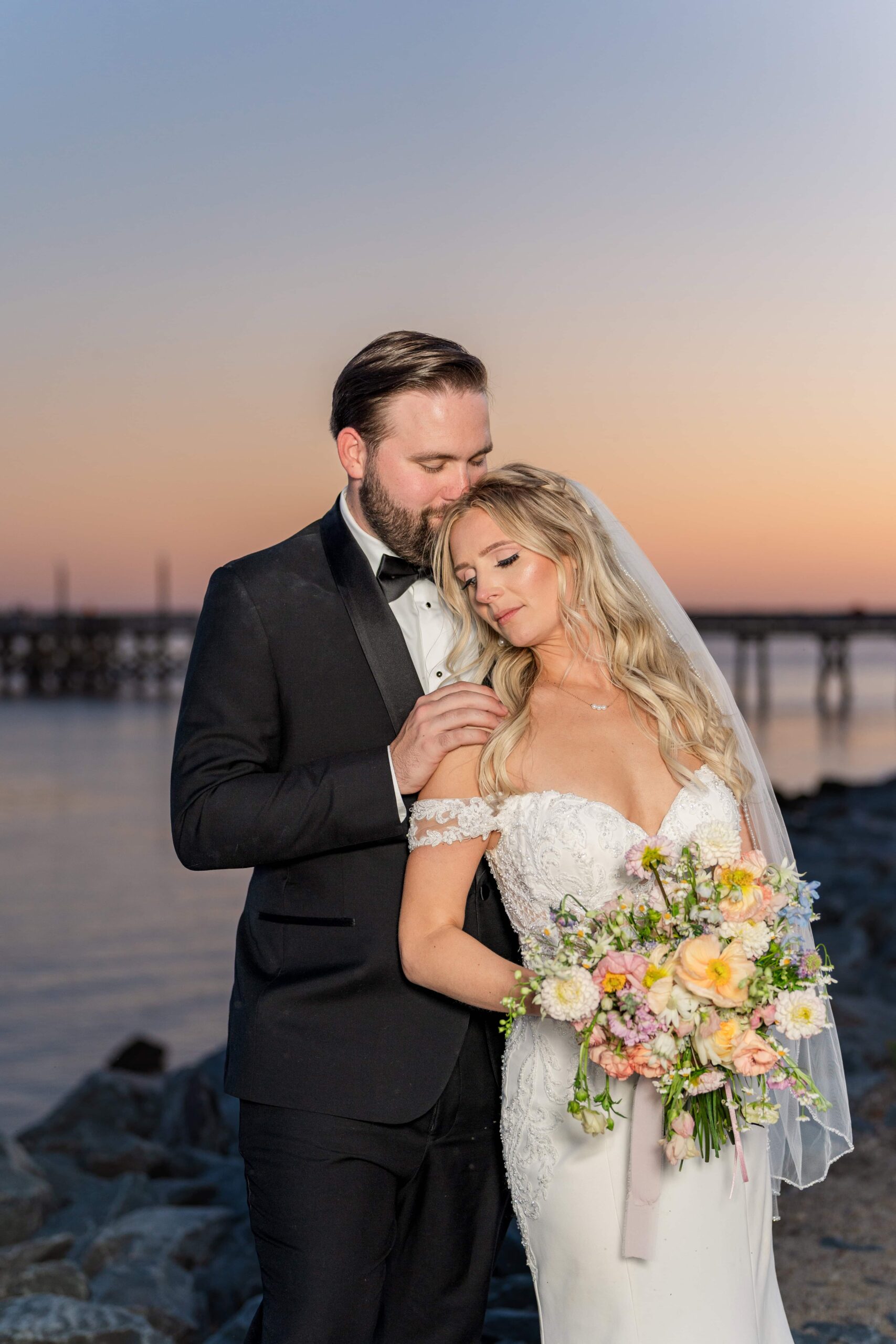 The bride and groom snuggle up together at the beach at sunset. The pier is visible off in the distance and the orange sunset reflects off of the ocean waves.