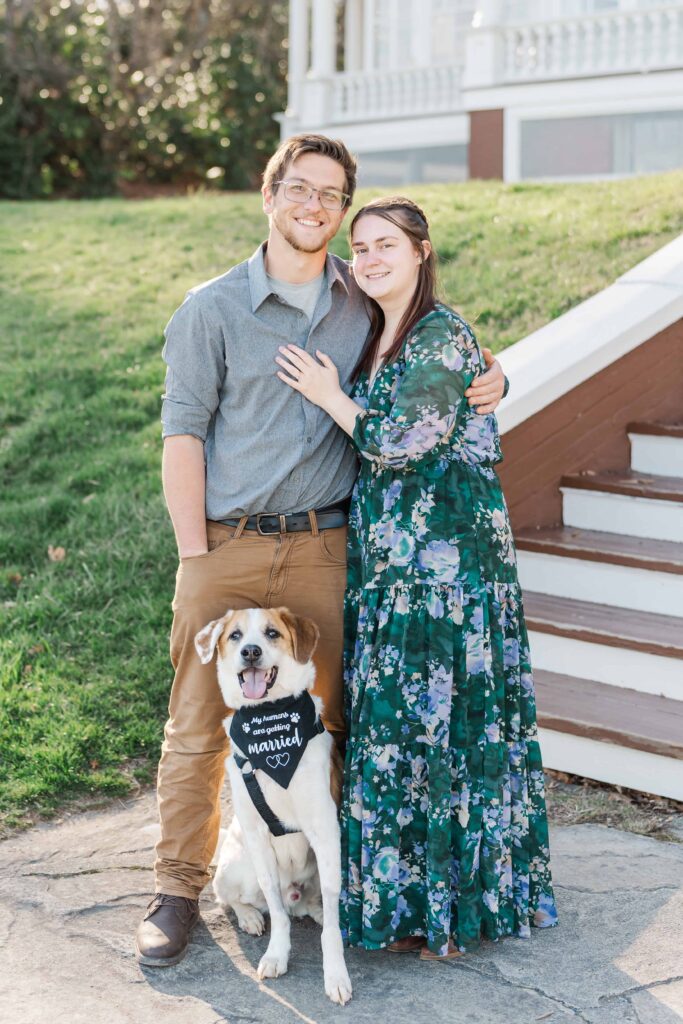 Couple smiles at Moses Cone Manor. They included their dog in their engagement session and Beau stands beside them with a bandana that reads "my humans are getting married"