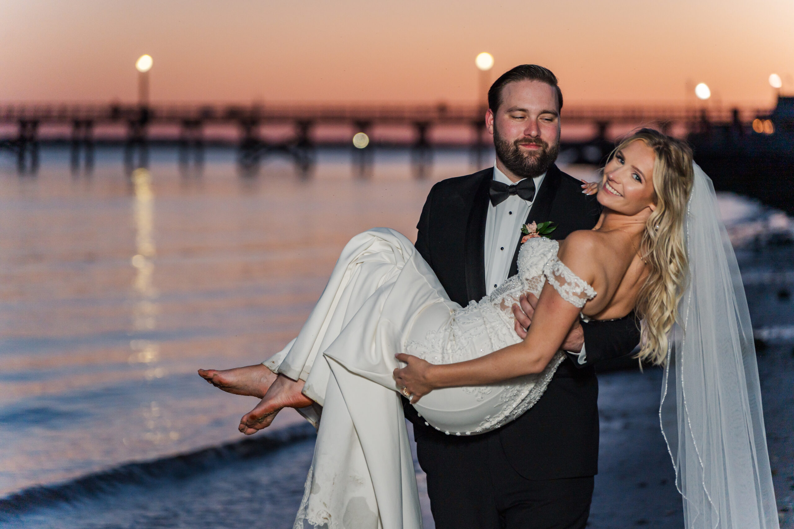 Groom carries his bride in his arms as he walks along the beach at sunset. The pier is visible off in the distance and the orange sunset reflects off of the ocean waves.