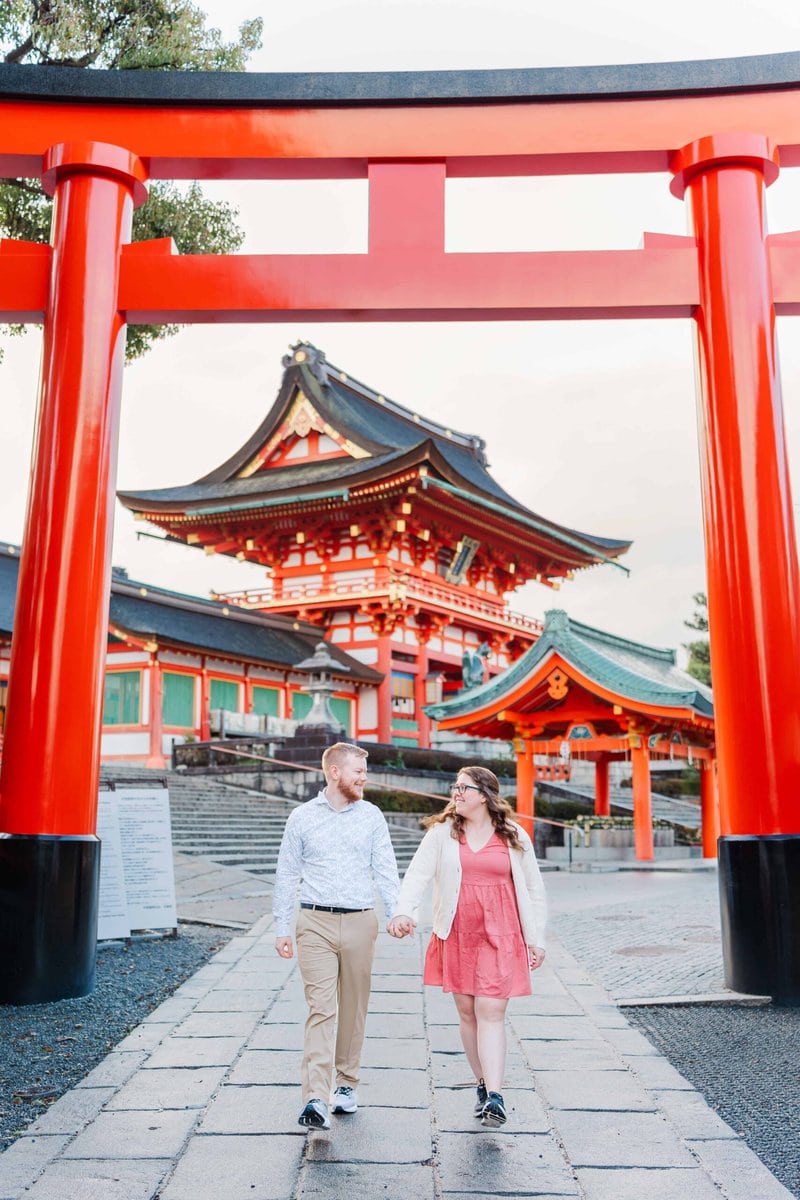 Couple walks underneath a bright red Torii Gate at Fushimi Inari Taisha in Kyoto, Japan.