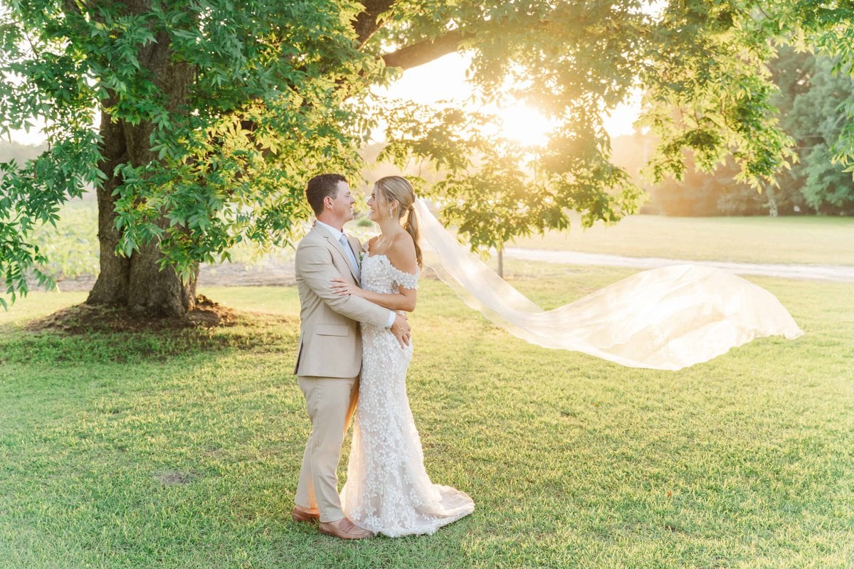 Maria and Dallon stand under a large tree for their wedding photos on their family farm. Maria's veil flows in the breeze and the sun sets behind them.