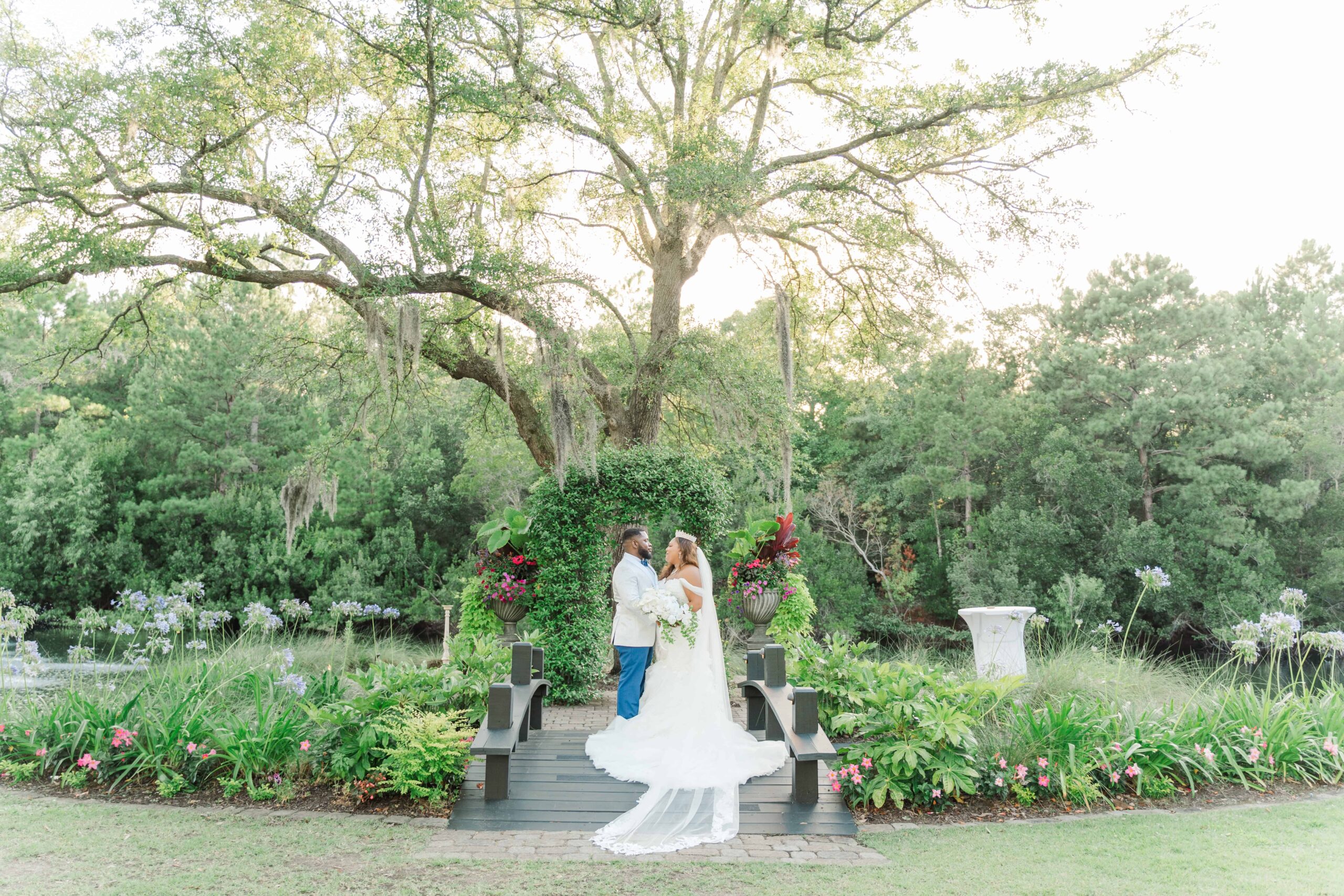 Kennedy and Joshua stand in the middle of the bridge that overlooks the pond at Hart Meadows Ranch. The evening sun glows around them, lighting up the oak tree draped with Spanish moss.