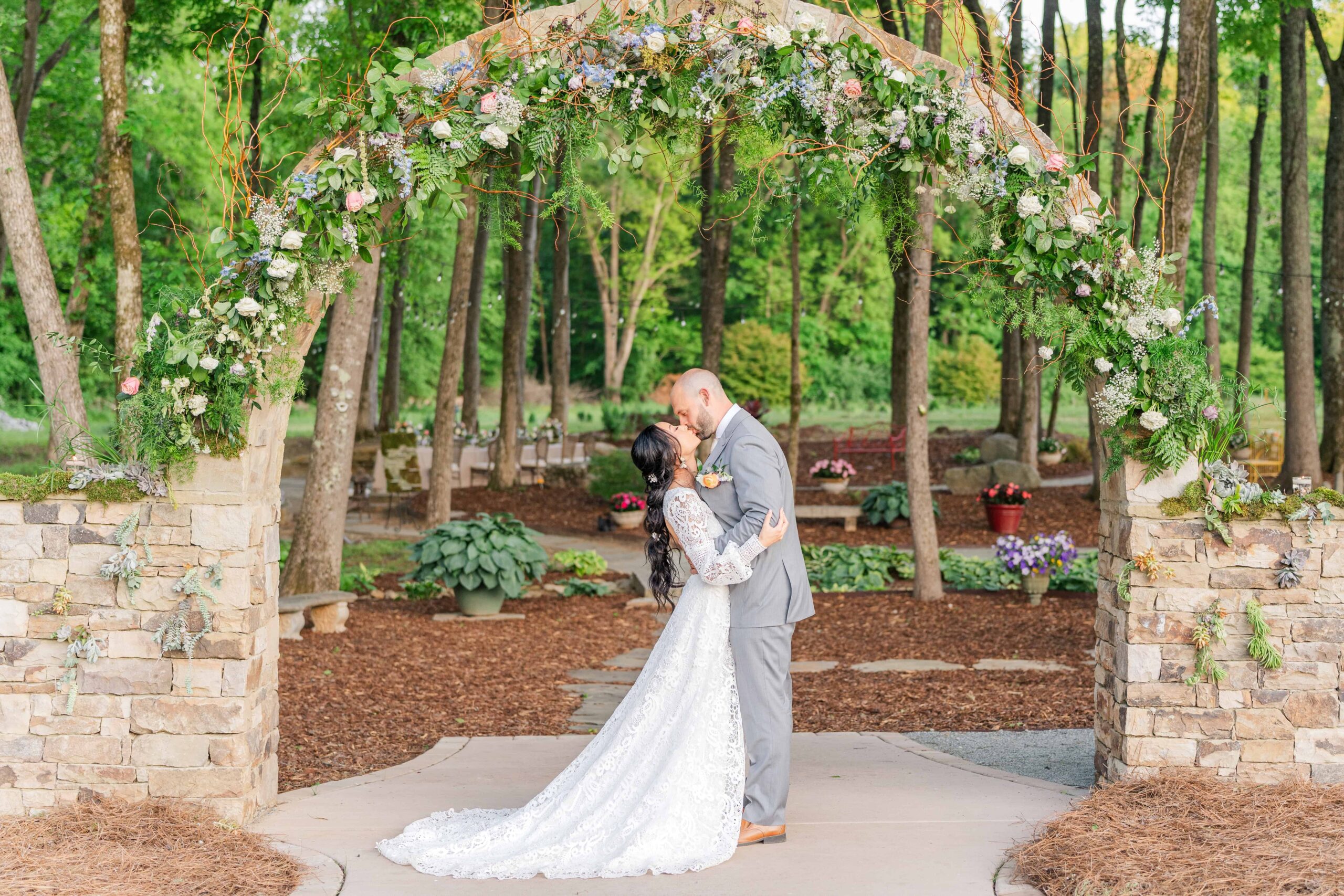 Couple shares a romantic kiss beneath a brick archway adorned with cascading florals, moss, and succulents at Low Meadows Estate.